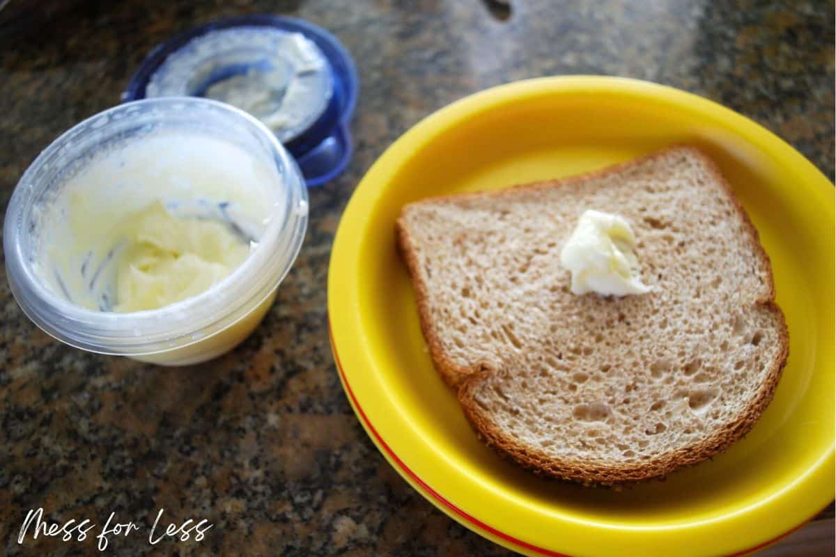 A slice of whole wheat bread with butter on a yellow plate, next to a container of more butter.