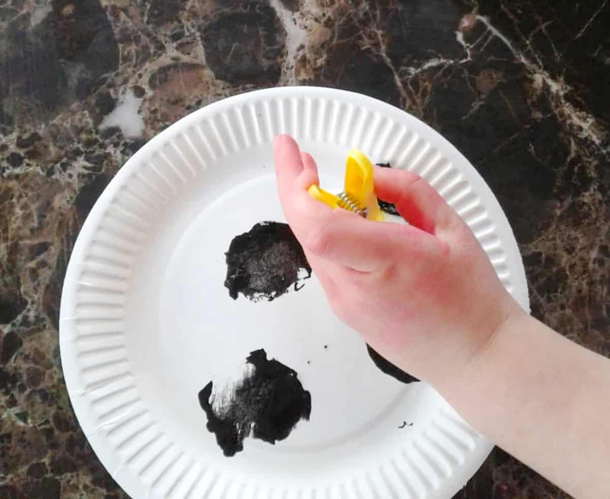 A hand holding a yellow clip presses it into black paint on a white paper plate, perfect for creating paper plate baby farm animals.