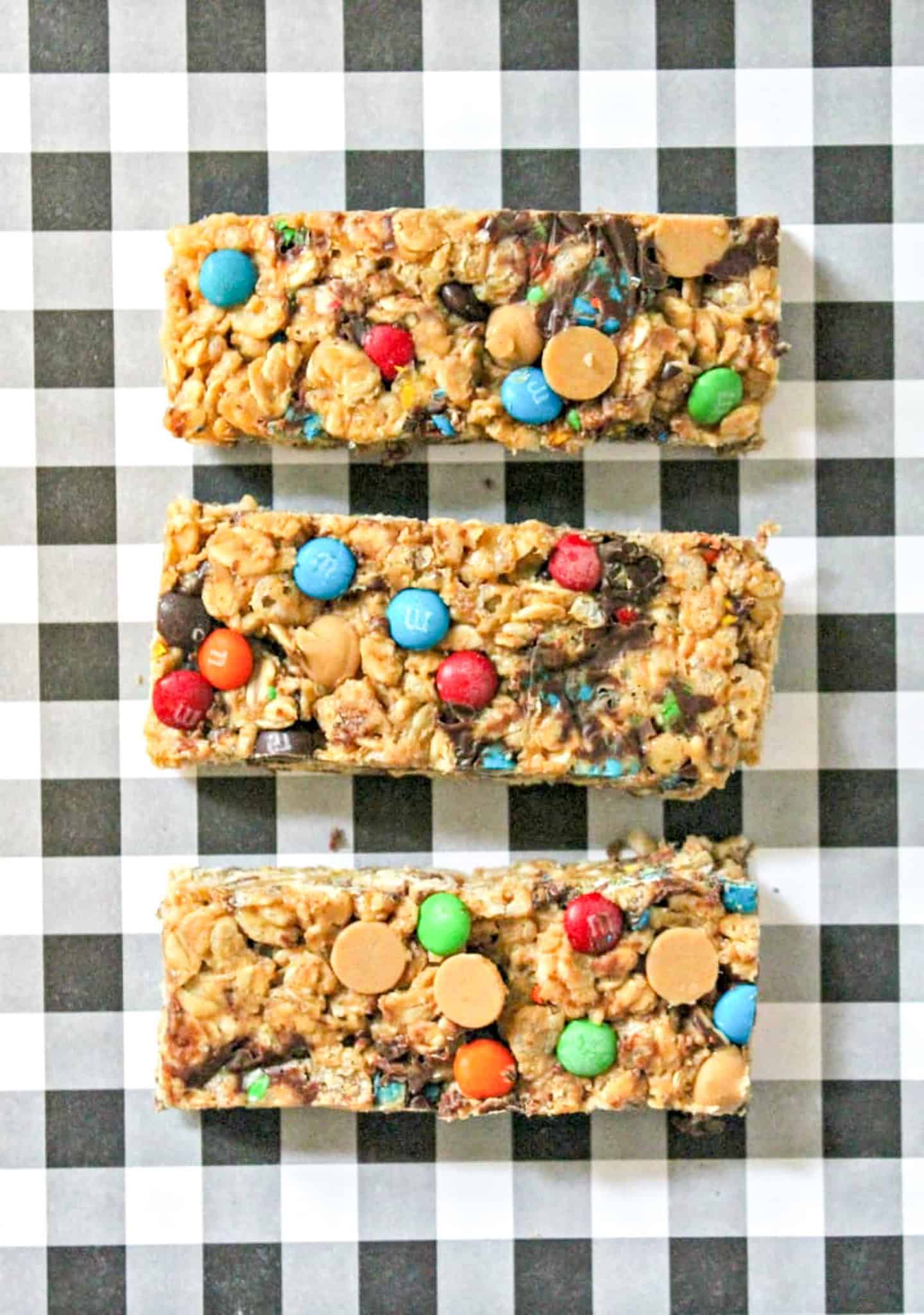 Three no bake granola bars with colorful candy pieces and chocolate chips are set on a black and white checkered background.