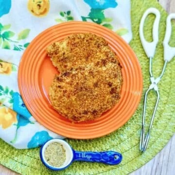 Breaded and baked pork chops on an orange plate with seasoning and tongs on a floral placemat
