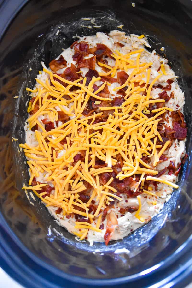 Crack Chicken topped with a generous layer of shredded cheddar cheese and crispy crumbled bacon in a slow cooker.
