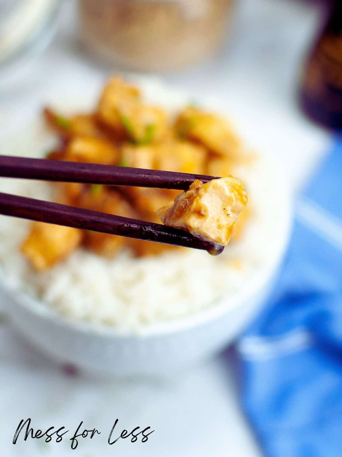 Chopsticks holding up piece of orange chicken over bowl of chicken and rice
