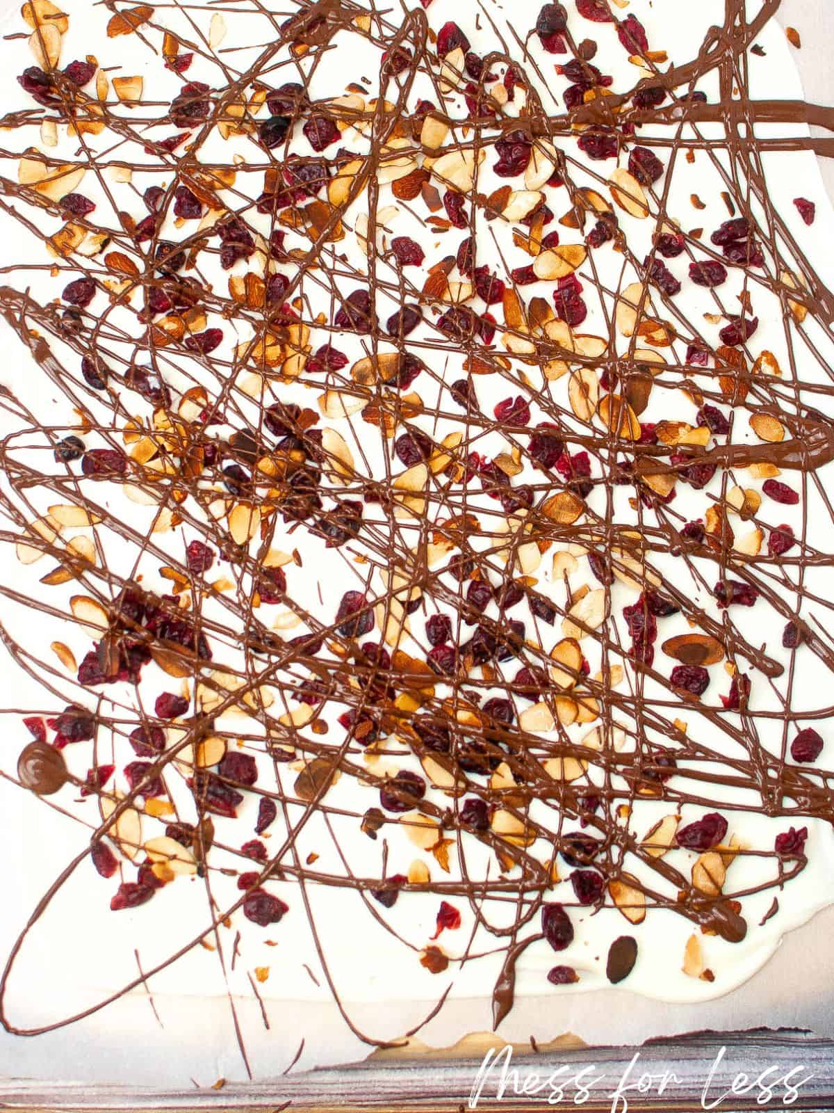 White chocolate spread on parchment paper with almonds craisins and chocolate drizzled on top