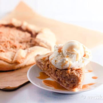 A slice of apple pie topped with a scoop of vanilla ice cream and drizzled with caramel sauce sits elegantly on a white plate. In the background, the rest of the pie rests on parchment paper, resembling a rustic crumb galette recipe.