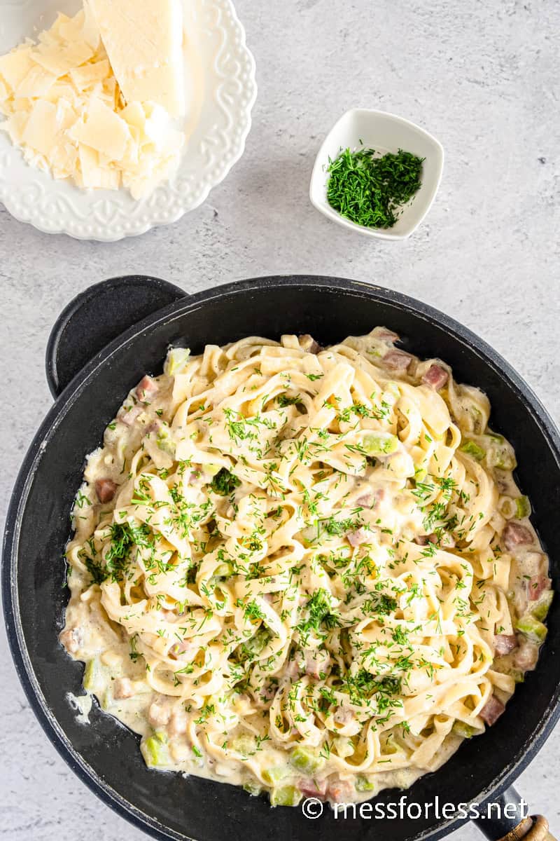A skillet with creamy pasta accented by crispy bacon and topped with chopped herbs. A small bowl of green herbs and a plate of shaved cheese are on the side. The background is a light, textured surface.