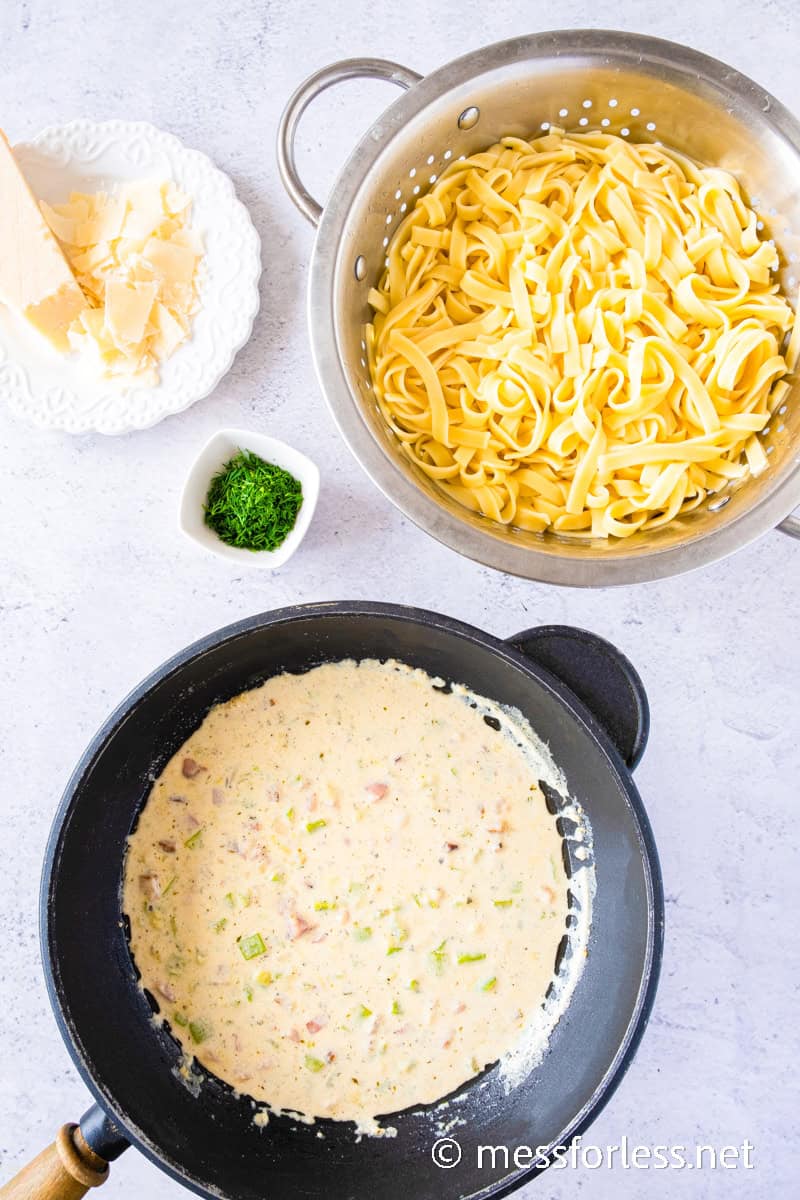 A colander with cooked fettuccine, a skillet containing a creamy pasta sauce blending bits of bacon and zucchini, and a plate with grated cheese next to a small bowl of chopped herbs on a light surface.