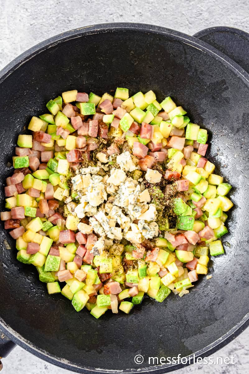 A black pan containing diced zucchini, crispy bacon, and crumbled blue cheese.