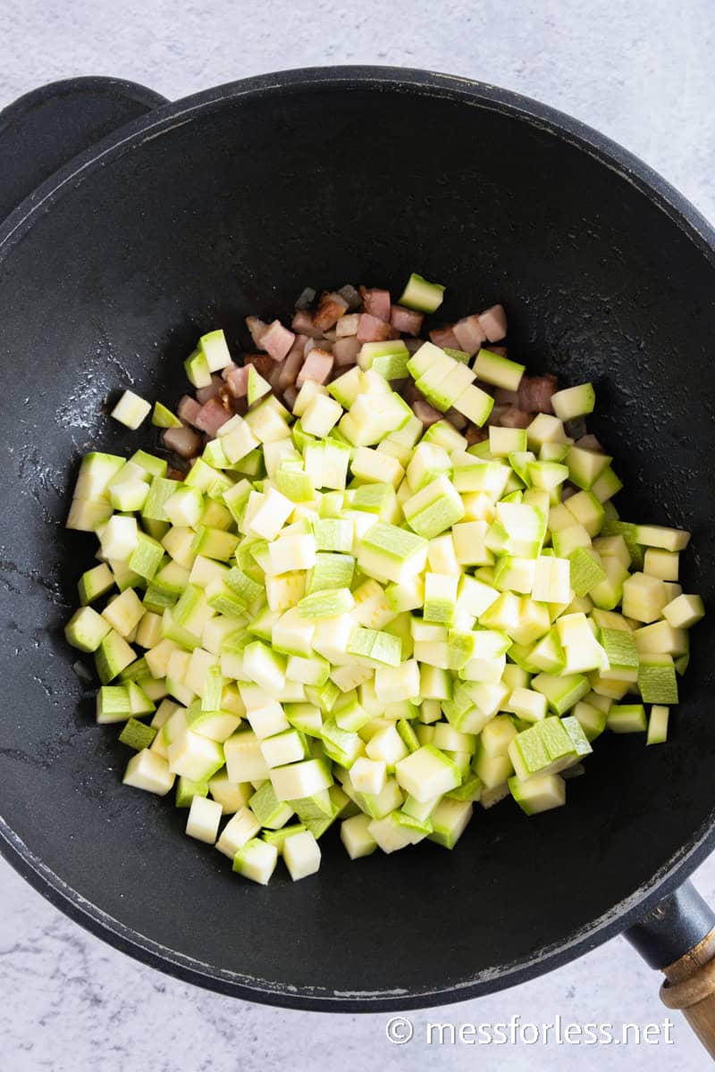 A frying pan on the stovetop contains chopped zucchini, diced onions, and bacon sizzling to perfection. The partially cooked vegetables create a tempting base for a creamy pasta dish.
