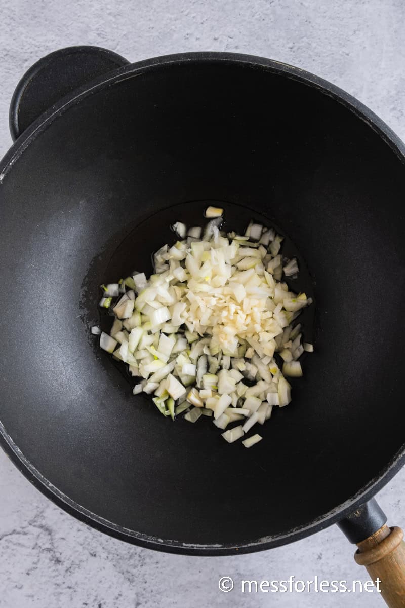 Chopped onions and garlic sizzle in a black frying pan with a wooden handle, set on a light gray surface.