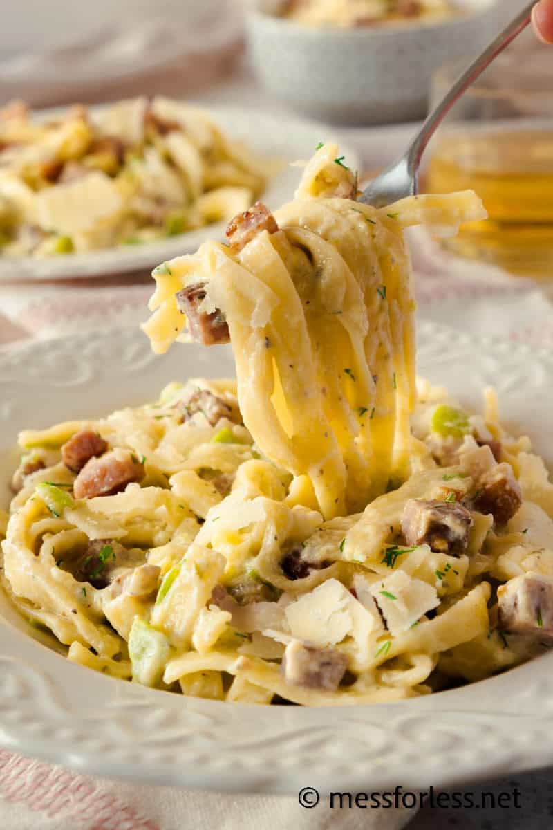 A plate of creamy fettuccine pasta with chunks of bacon and tender zucchini, sprinkled with herbs, is being twirled by a fork. The dish appears rich and hearty, served in a decorative white bowl. A glass of amber-colored beverage sits in the background.