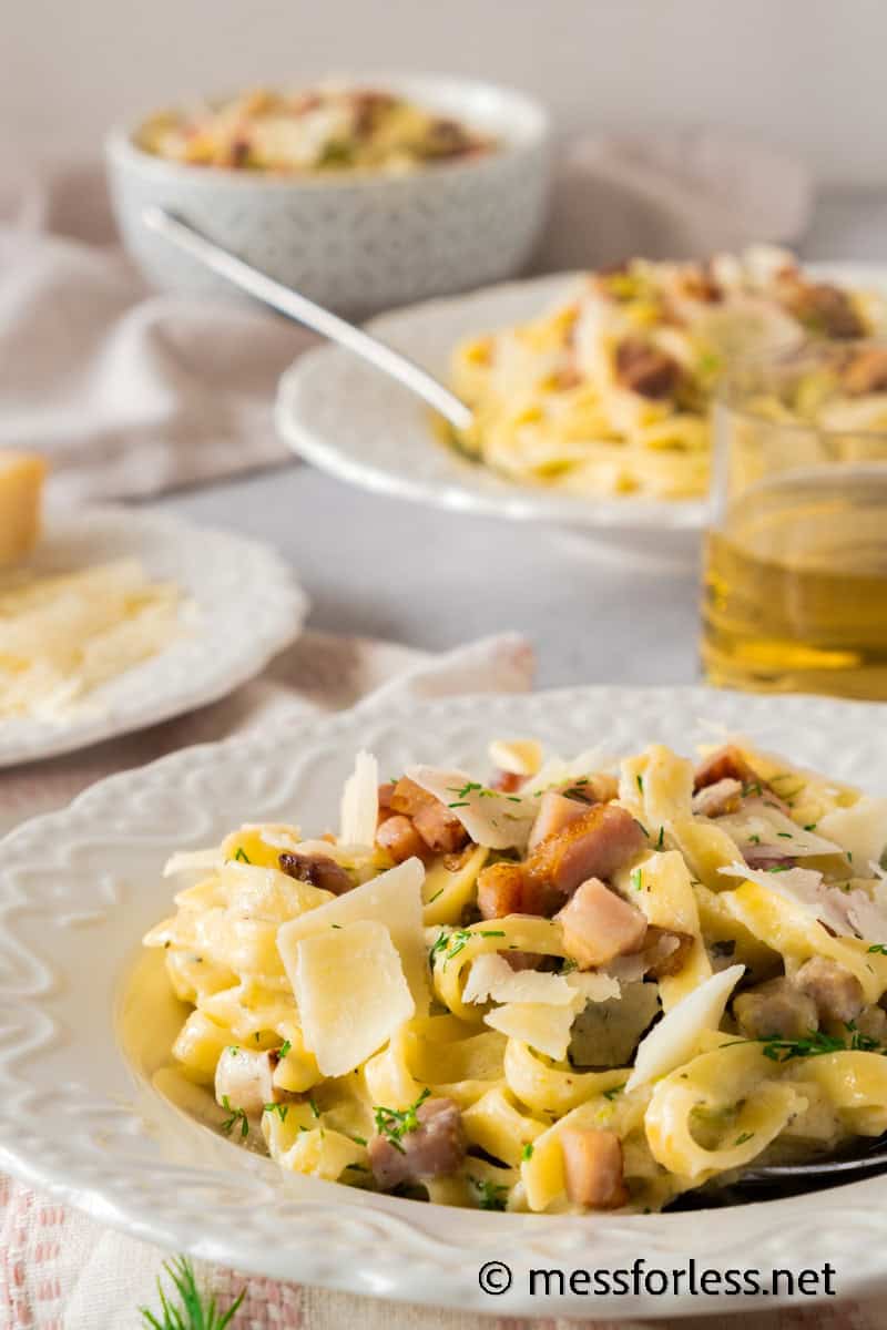 A close-up of a plate of creamy pasta, enriched with chunks of bacon and shaved cheese, alongside zucchini. Another plate is visible in the background, paired with a small glass of drink and a wedge of cheese, all set on a light-colored tablecloth.