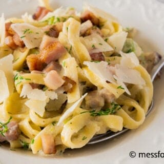 A close-up of creamy pasta topped with crispy bacon pieces, thin slices of zucchini, and shaved cheese, garnished with fresh herbs. A fork rests on the side of the white plate.
