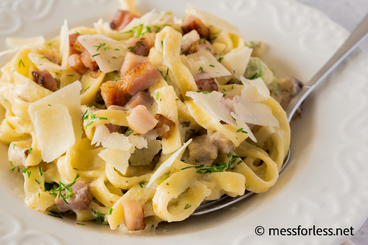A close-up of creamy pasta topped with crispy bacon pieces, thin slices of zucchini, and shaved cheese, garnished with fresh herbs. A fork rests on the side of the white plate.