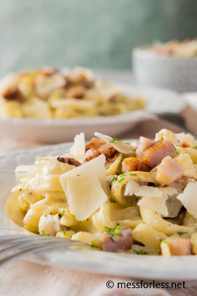 A close-up of creamy fettuccine pasta topped with crispy bacon pieces and Parmesan shavings. The rich sauce perfectly coats the strands, with herbs sprinkled on top. Thin slices of zucchini add color, while another dish of pasta is blurred in the background.