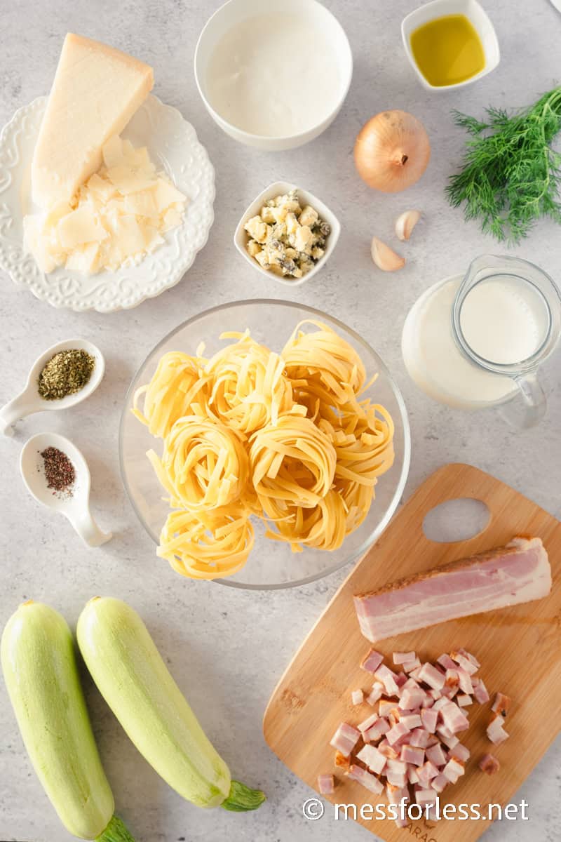 Ingredients laid out on a countertop for creamy pasta preparation include Parmesan cheese, tagliatelle nests, olive oil, onion, garlic, cream, zucchini, cubed bacon, herbs, and pepper.