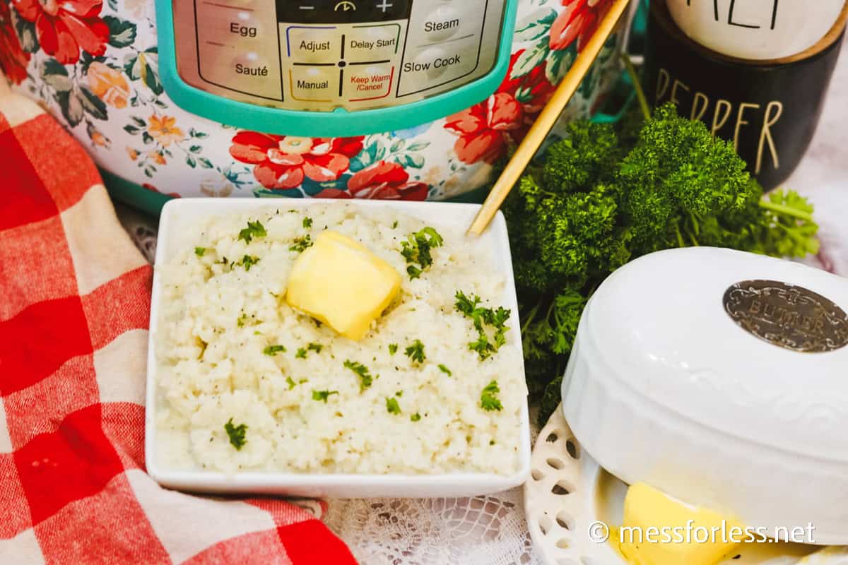 A bowl of mashed cauliflower rice topped with a pat of butter and garnished with parsley sits in front of a patterned Instant Pot. A red-striped cloth and butter dish are nearby, completing this delightful recipe scene.
