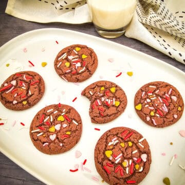 A white tray with six chocolate cookies made from Valentines Chocolate Cake Mix, adorned with red, pink, and yellow sprinkles. One cookie has a bite taken out. In the background, theres a glass of milk and a white cloth with black accents.