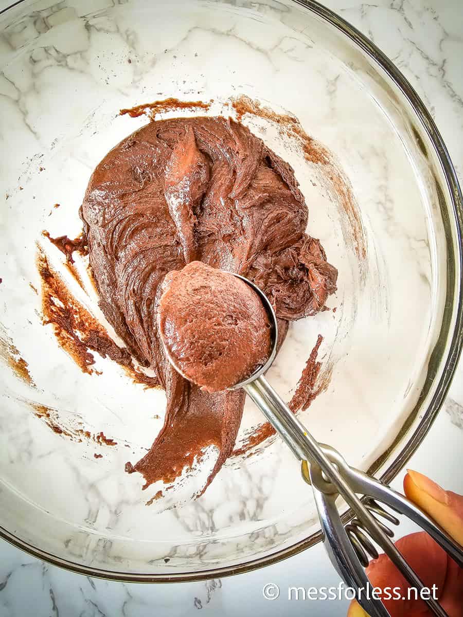 A glass bowl brimming with chocolate cookie dough for Valentines Chocolate Cake Mix Cookies rests on a marble countertop. A cookie scoop is poised to form perfect portions. The image features a watermark from messforless.net at the bottom right corner.