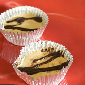 Two peanut butter cups in foil wrappers, topped with dark chocolate drizzle, on a red surface—perfect for a homemade treat inspired by a Weight Watchers peanut butter cups recipe.