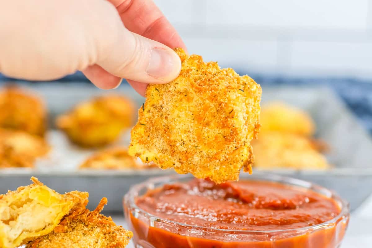 A hand holds a crispy, air fryer ravioli dipped in marinara sauce, with more golden delights resting on a tray in the background.