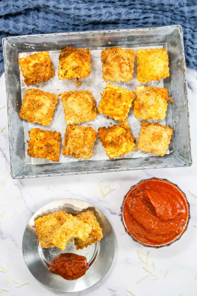 Air fryer ravioli recipe: Crispy fried ravioli on a tray with a bowl of marinara sauce, set on a marble surface with a blue textured cloth.