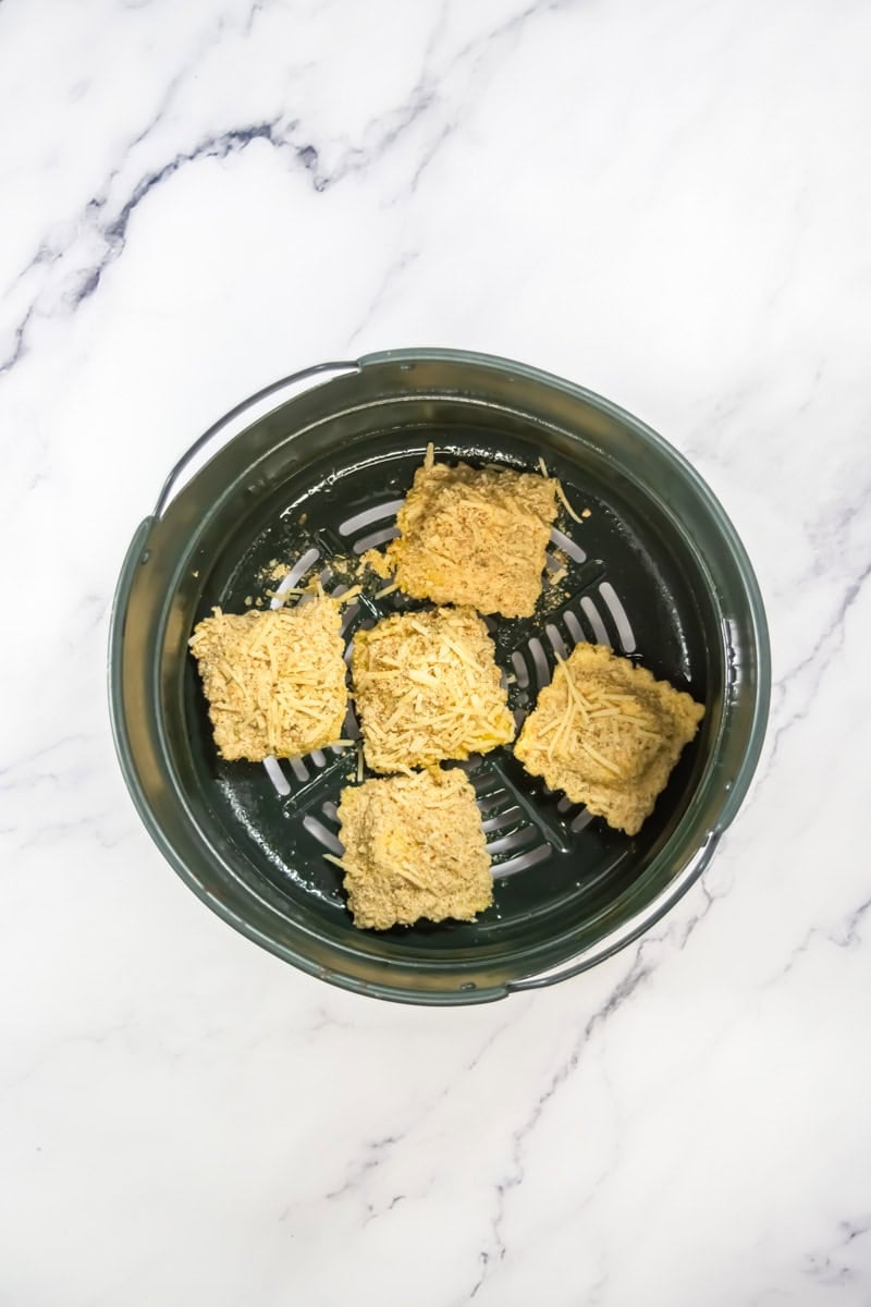 Four golden-brown pieces of breaded ravioli in a green air fryer basket on a marble surface.