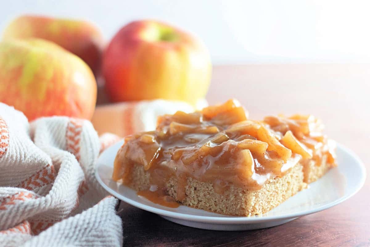 A delightful slice of caramel apple cake sits on a white plate, its rich caramel topping glistening invitingly. Surrounded by fresh apples and a striped cloth, this tempting treat hints at the delicious recipe behind its creation.