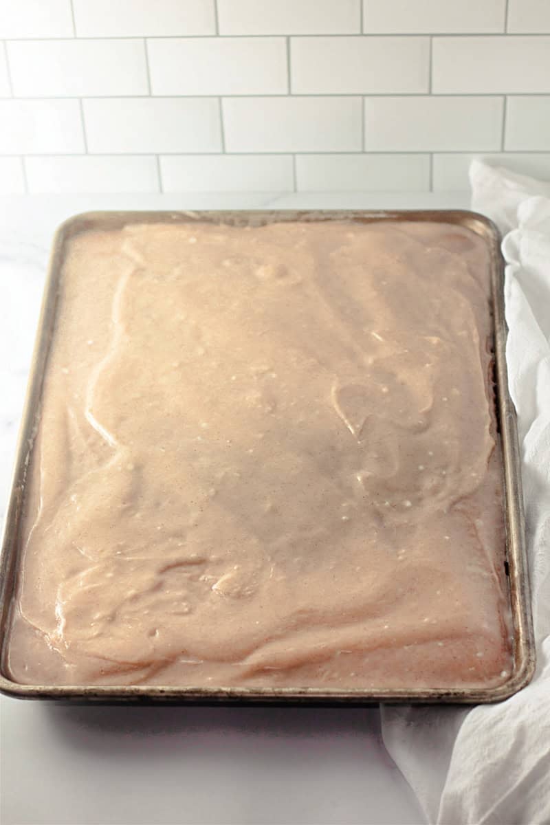 A sheet pan filled with smooth, light brown caramel cake batter rests against a white tiled background, promising a sweet and indulgent treat.