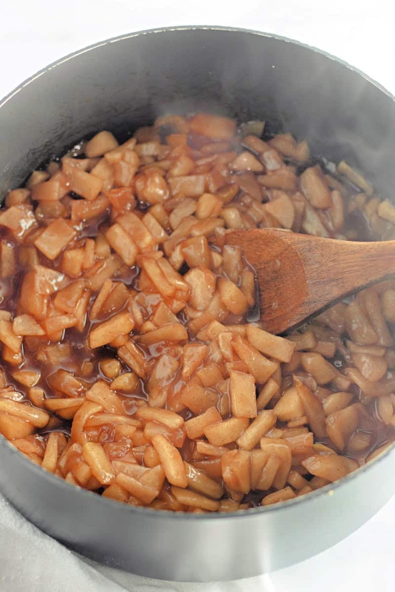 A pot of diced apples simmering with cinnamon, gently stirred with a wooden spoon. Steam rises as the aroma evokes thoughts of a caramel apple cake recipe in the making.