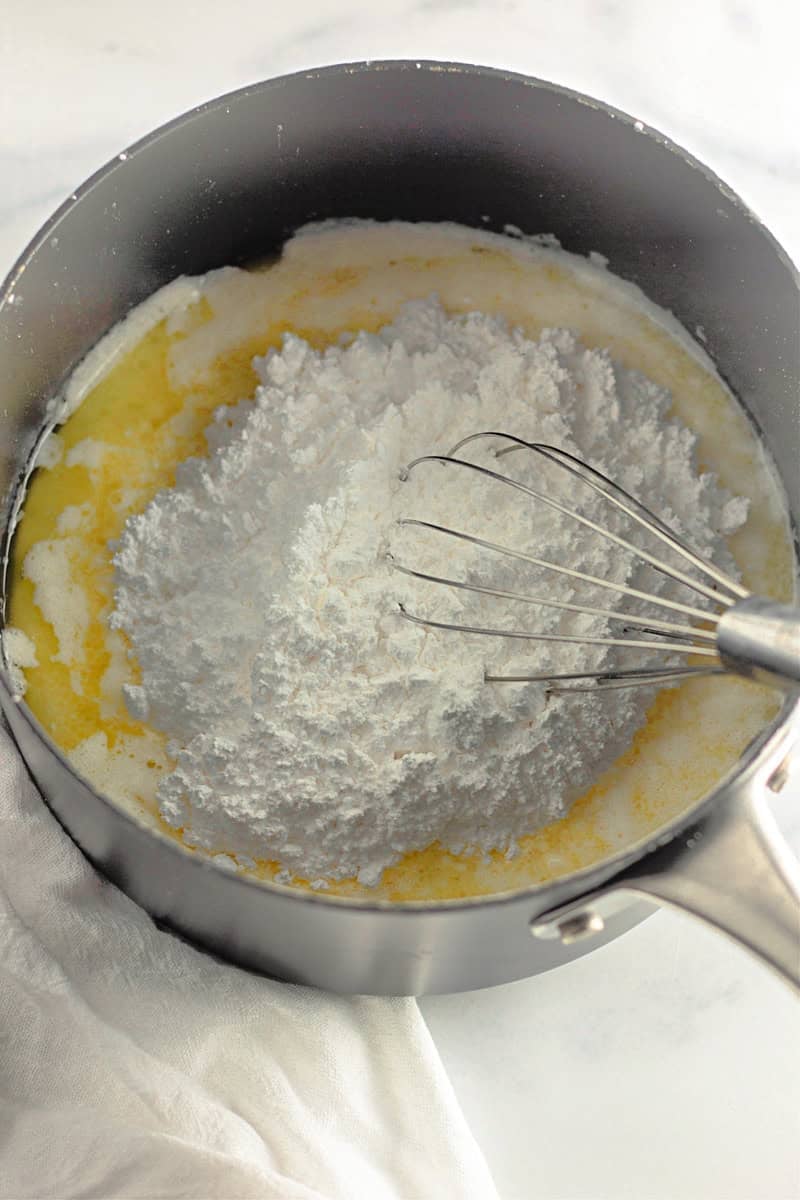 Powdered sugar and melted butter meld together like a silky caramel, as they swirl in a saucepan with a whisk on a light surface, laying the foundation for an unforgettable apple-inspired cake recipe.
