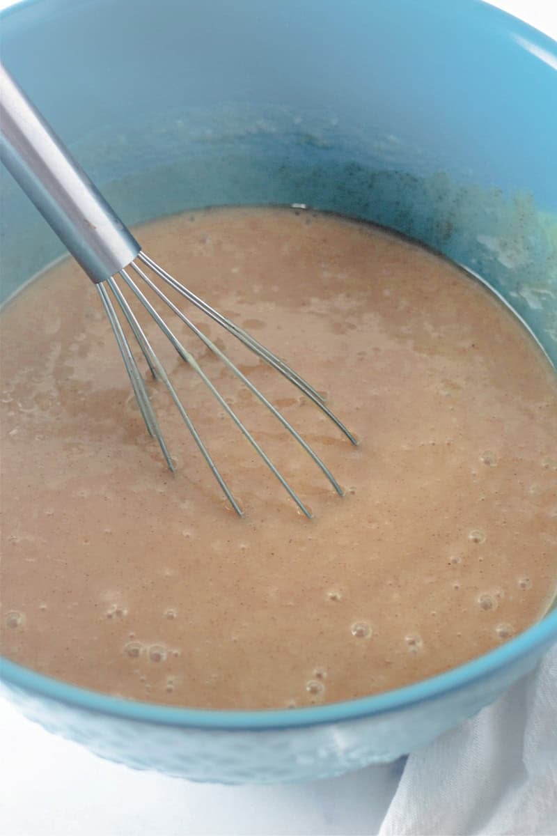 A blue bowl cradles the beige batter for a caramel apple cake, being expertly mixed with a metal whisk.