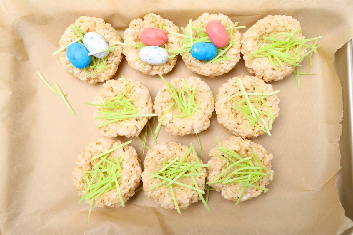 Easter nest Rice Krispie treats in progress—partially decorated with green edible grass and candy eggs, placed on a parchment-lined baking tray.