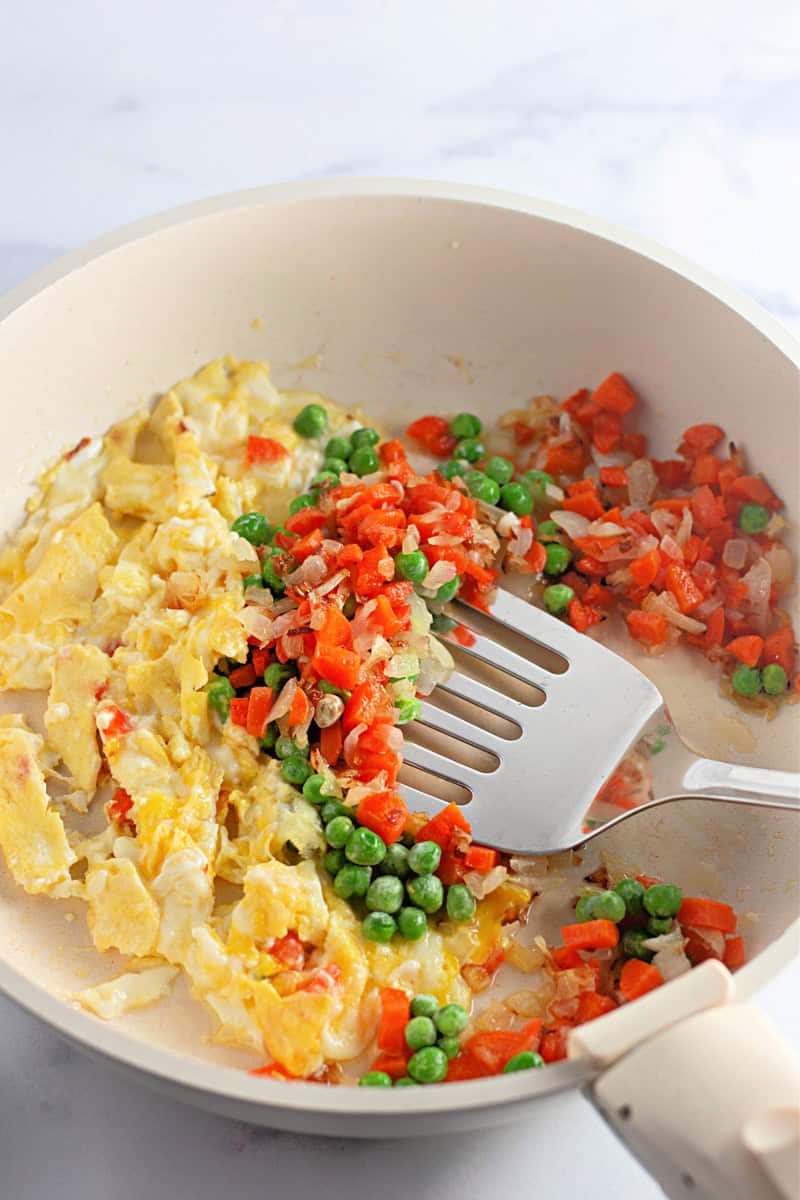 An easy recipe unfolds as eggs, peas, and diced carrots sizzle in a white frying pan with a spatula—a delightful prelude to homemade fried rice.