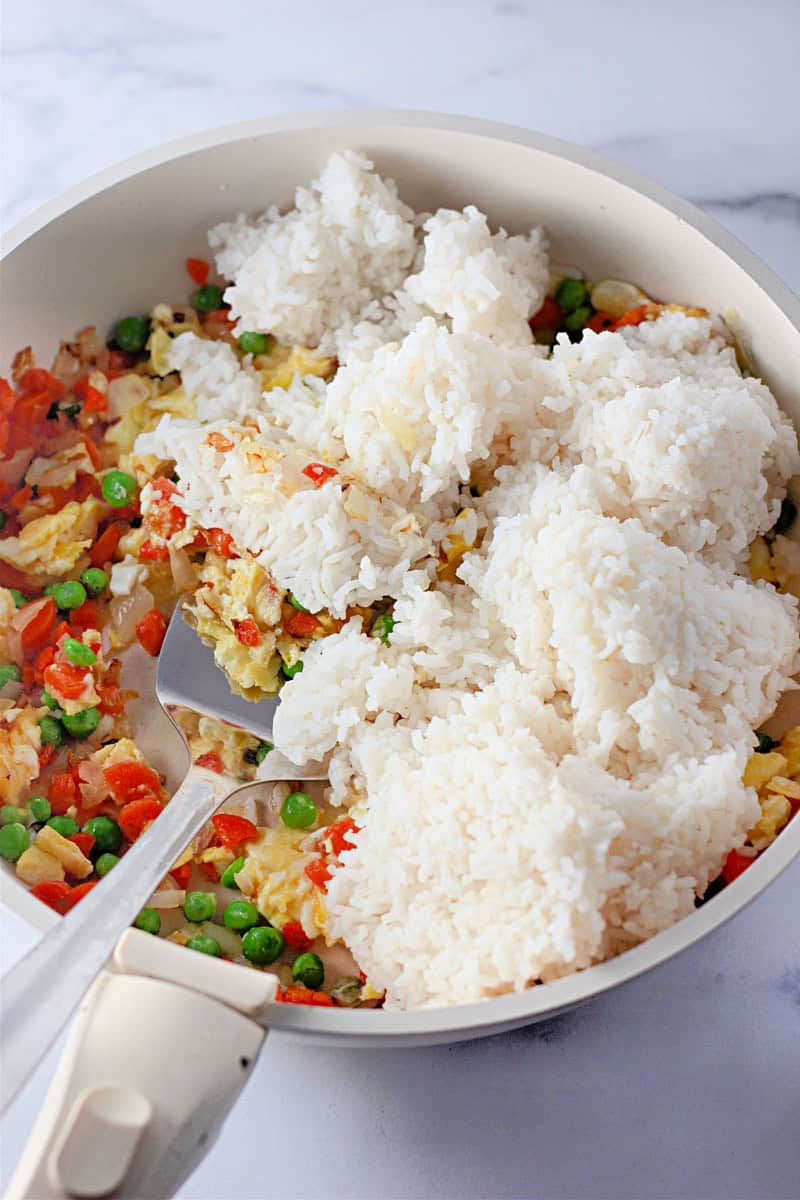 A frying pan sizzles with an easy fried rice recipe, featuring rice, peas, diced bell peppers, and eggs cooking to perfection on a white surface.