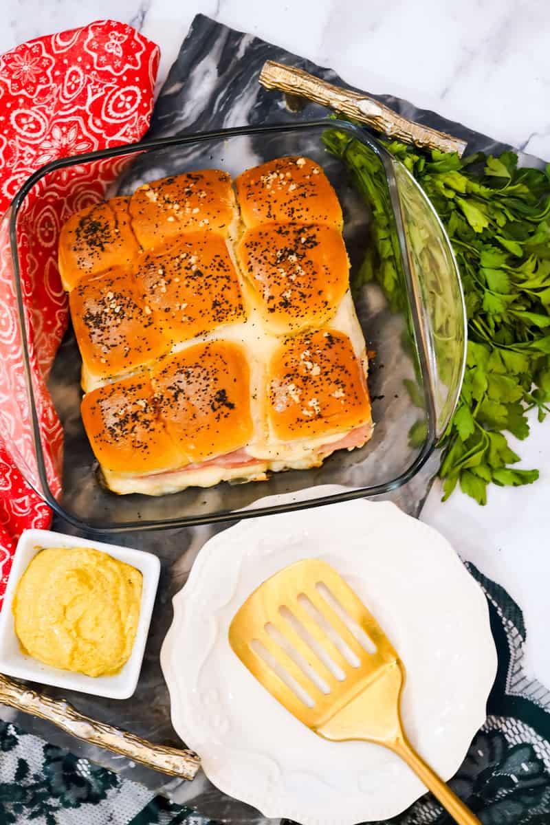 Baked sliders with Swiss cheese and ham in a glass dish, garnished with poppy seeds, parsley, and a touch of mustard. A spatula rests nearby on the marble background.