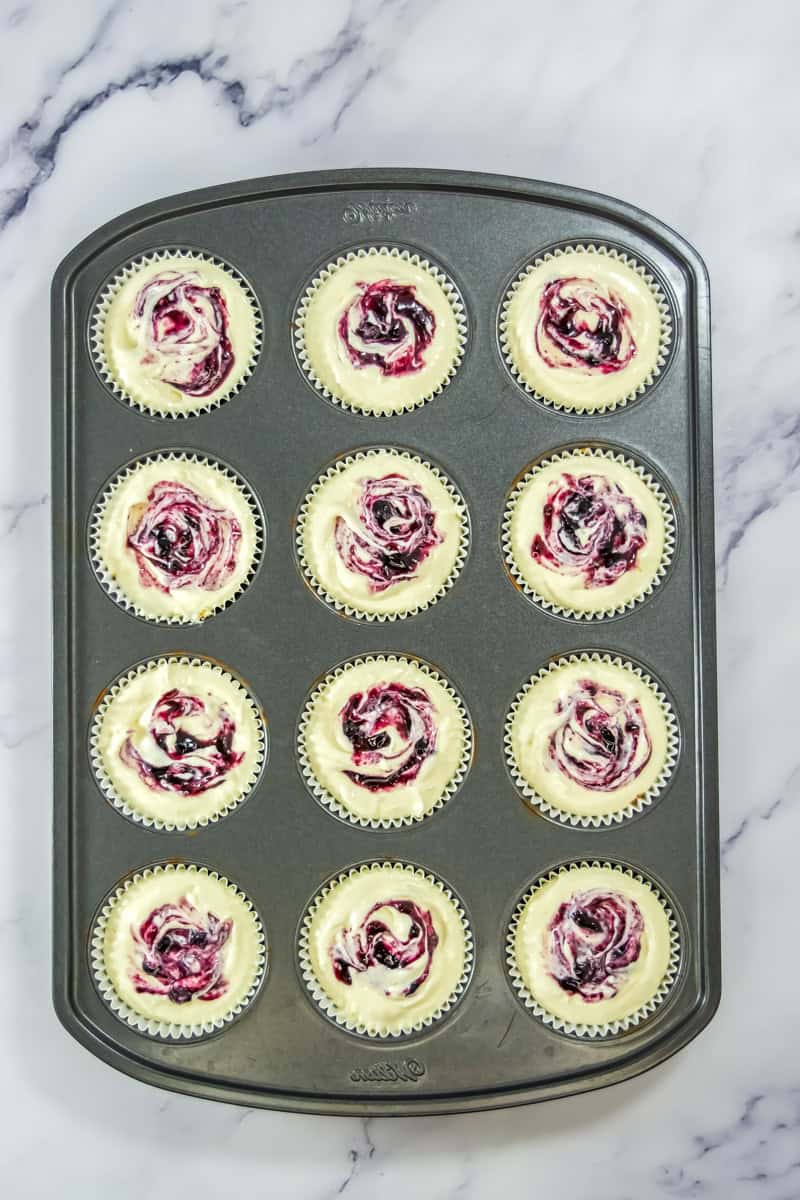 Muffin tin with 12 mini Lemon Blueberry Cheesecakes, each cupcake topped with a vibrant purple berry swirl, displayed on a marble surface.