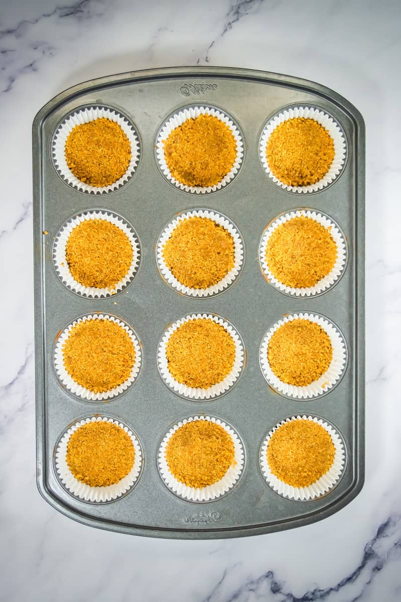 A muffin tin with 12 cupcake liners, each filled with a golden graham cracker crust, perfect for creating mini cheesecake treats topped with blueberry or lemon flavors.