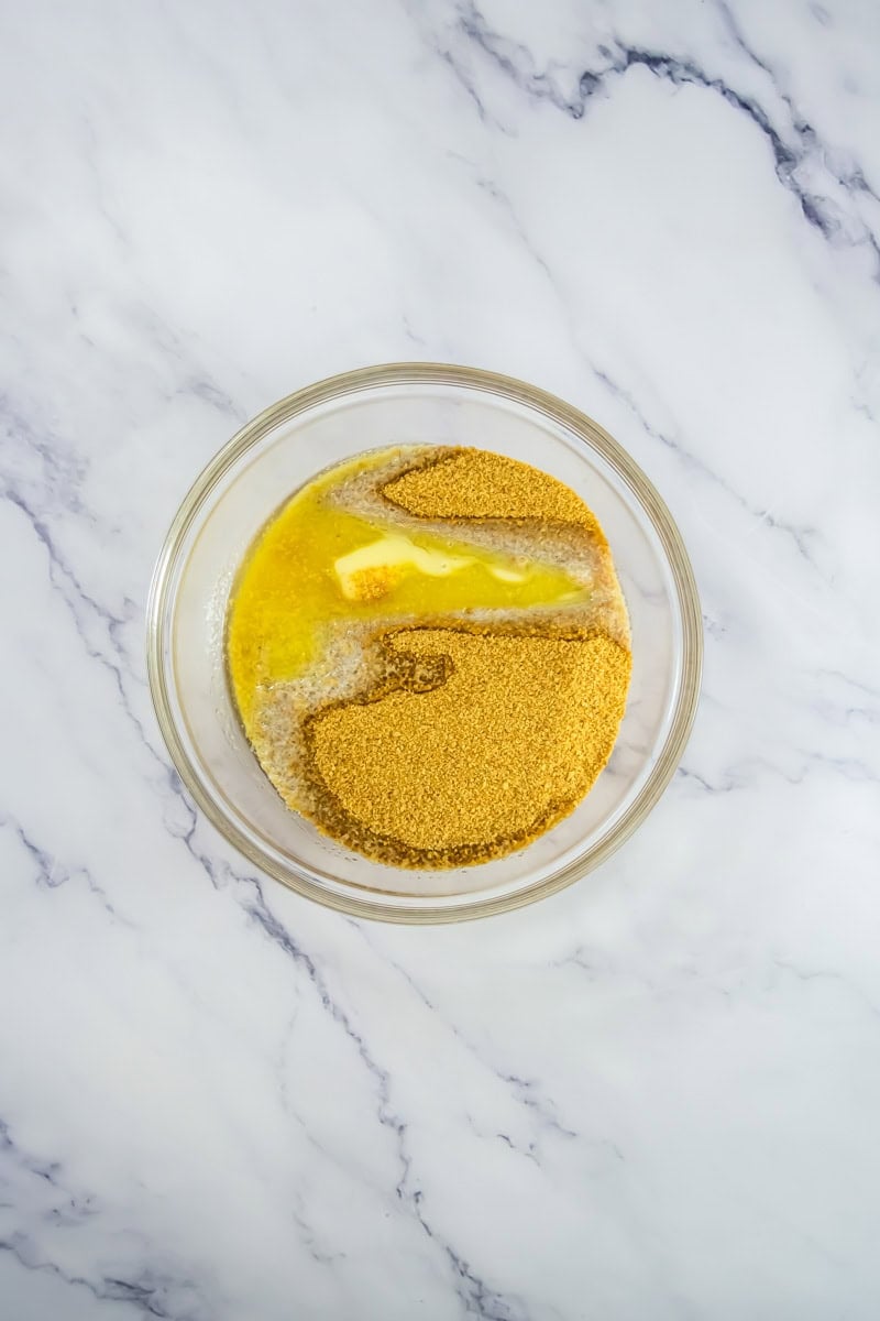 A glass bowl with melted butter and graham cracker crumbs on a white marble surface—perfect for creating Lemon Blueberry Mini Cheesecake.