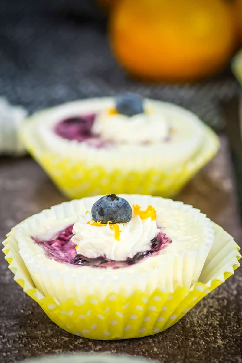 Mini cheesecake in a yellow polka dot cup, topped with whipped cream, fresh blueberry, and a hint of orange zest—this lemon blueberry mini cheesecake is the perfect sweet bite.