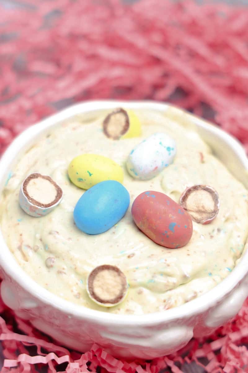 A bowl of creamy Robins Egg Easter Dip, topped with colorful candy eggs, sits elegantly on a pink shredded paper background.