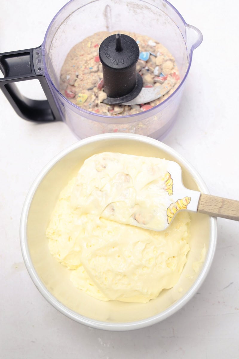 Creating a Robins Egg Easter Dip involves mixing crushed cookies with cream in a bowl next to a food processor on a white background.
