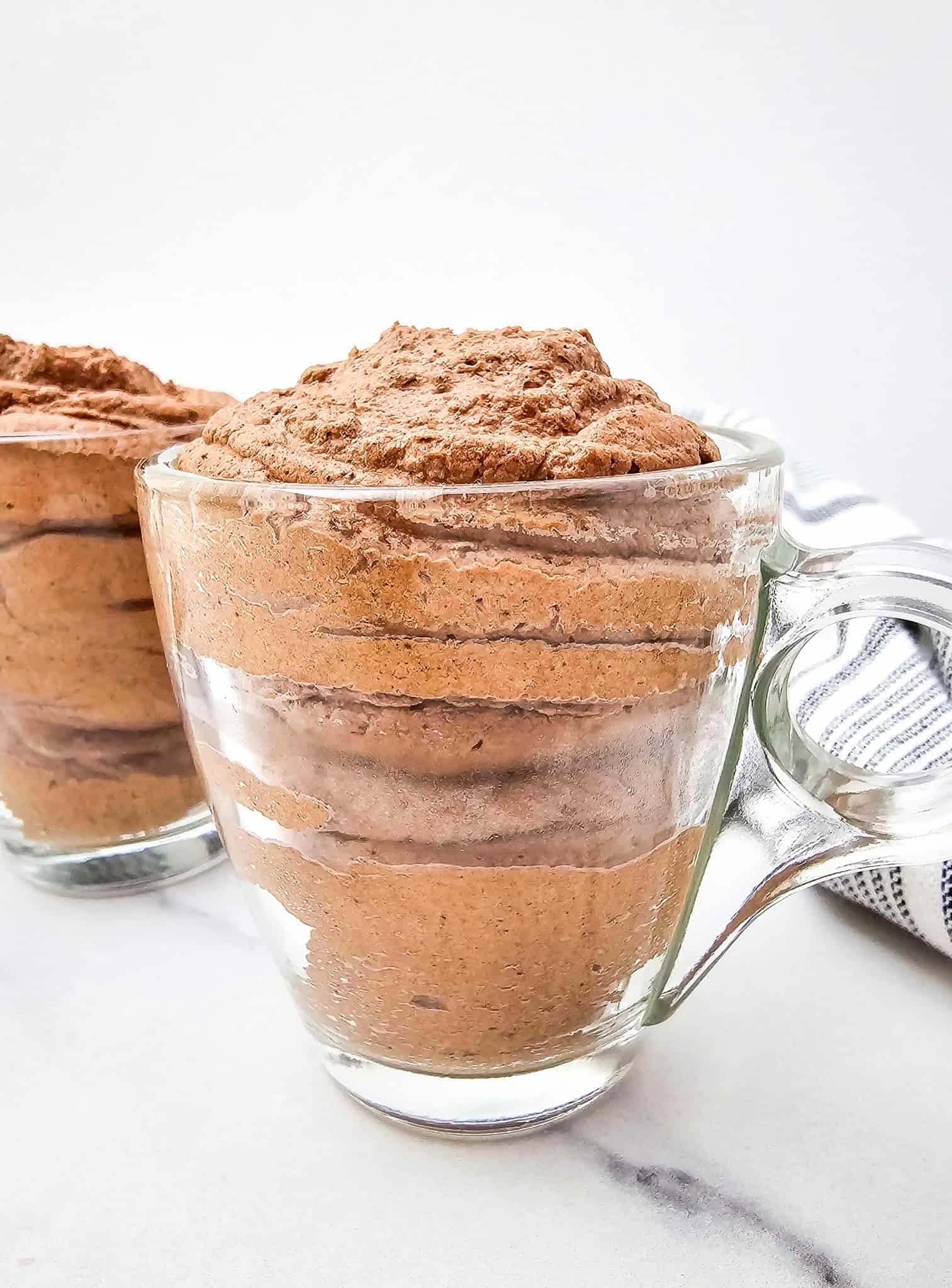 Glass mugs filled with 3 ingredient chocolate mousse sit on a white surface with a striped cloth nearby—perfect for a rich and creamy keto treat.