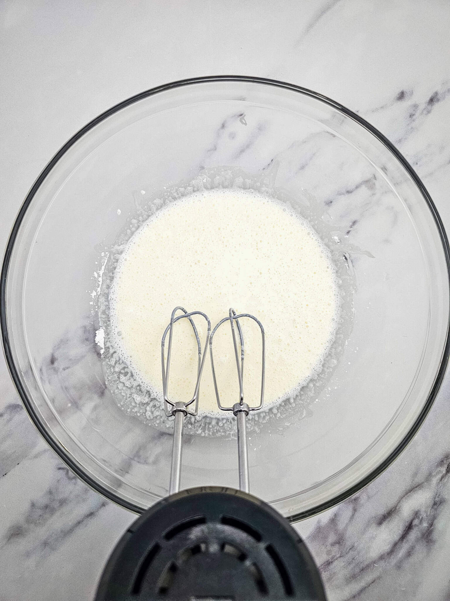 Glass bowl with white liquid and electric mixer beaters on a marble surface, viewed from above—perfect for whipping up a 3 ingredient keto dessert like Keto chocolate mousse.