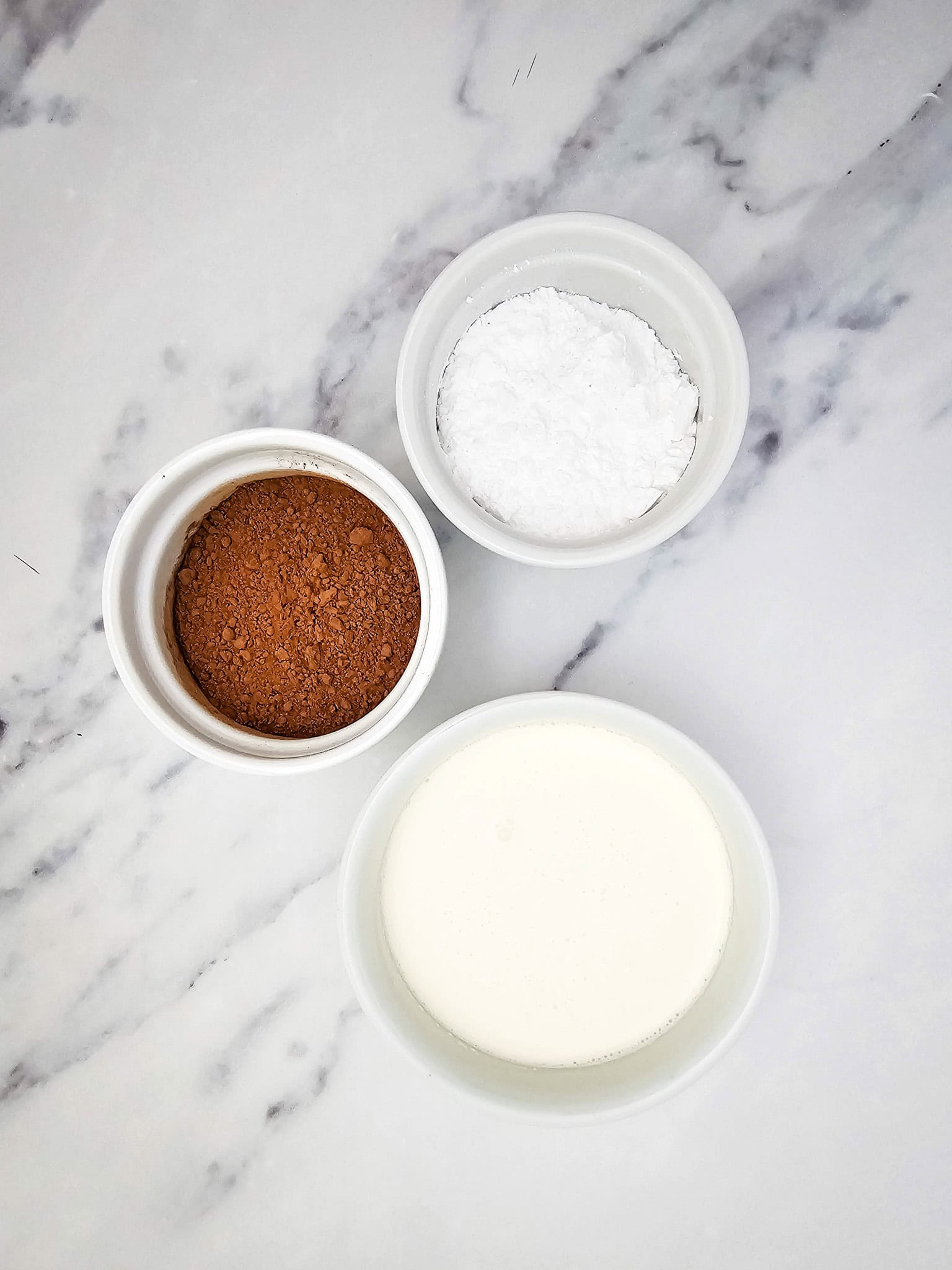 Three white bowls containing cocoa powder, powdered sugar, and cream on a marble surface—perfect for making a simple 3 ingredient Chocolate Mousse or keto dessert.