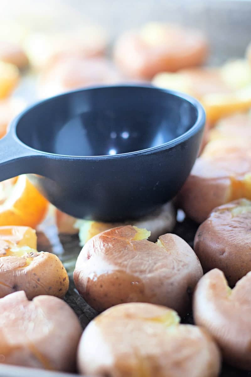 A black measuring cup presses down on cooked baby potatoes to flatten them, creating perfect smashed potatoes ready for a Garlic Parmesan finish.