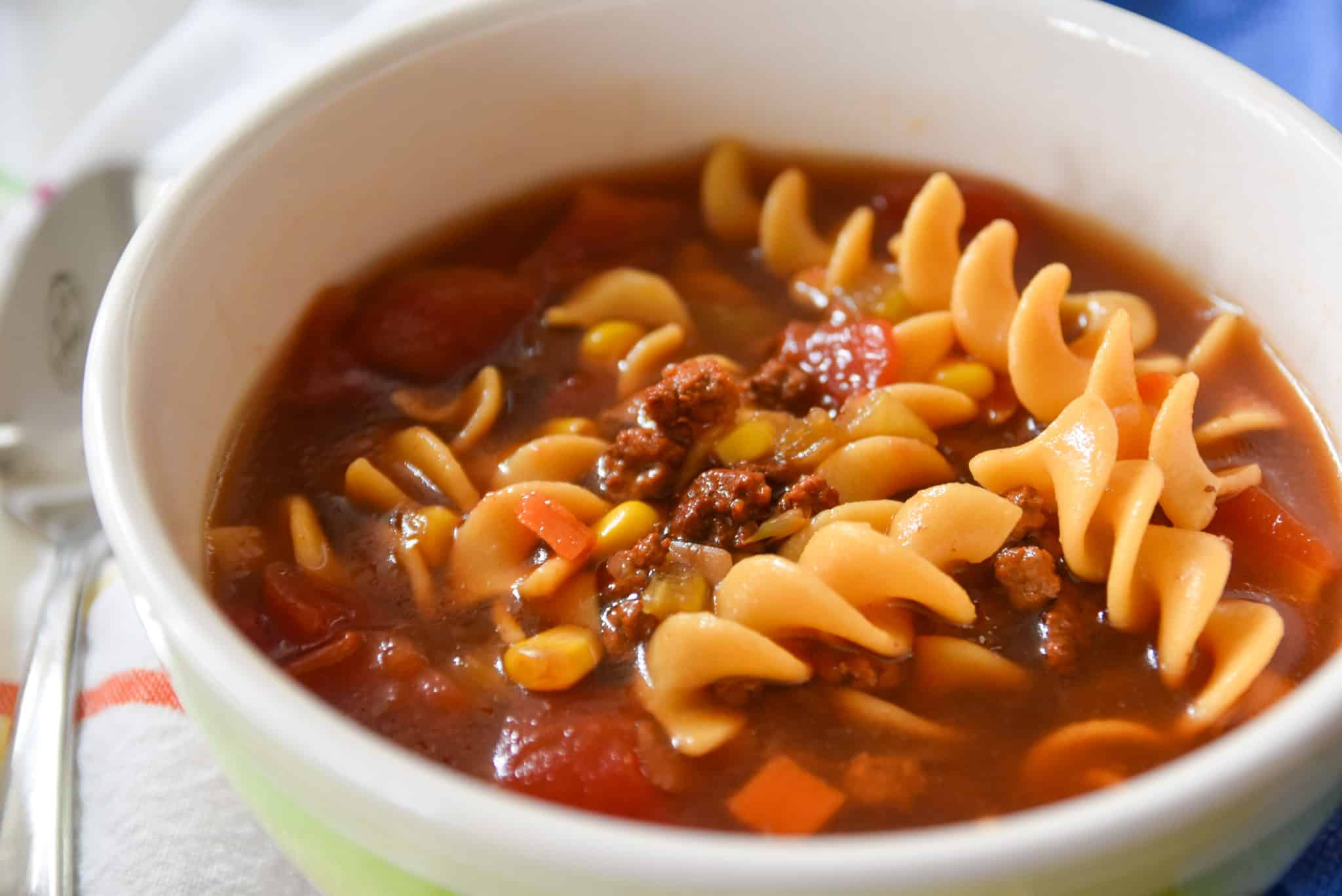 A comforting bowl of beef soup featuring a rich tomato base, spiral pasta, ground beef, corn, and diced vegetables.