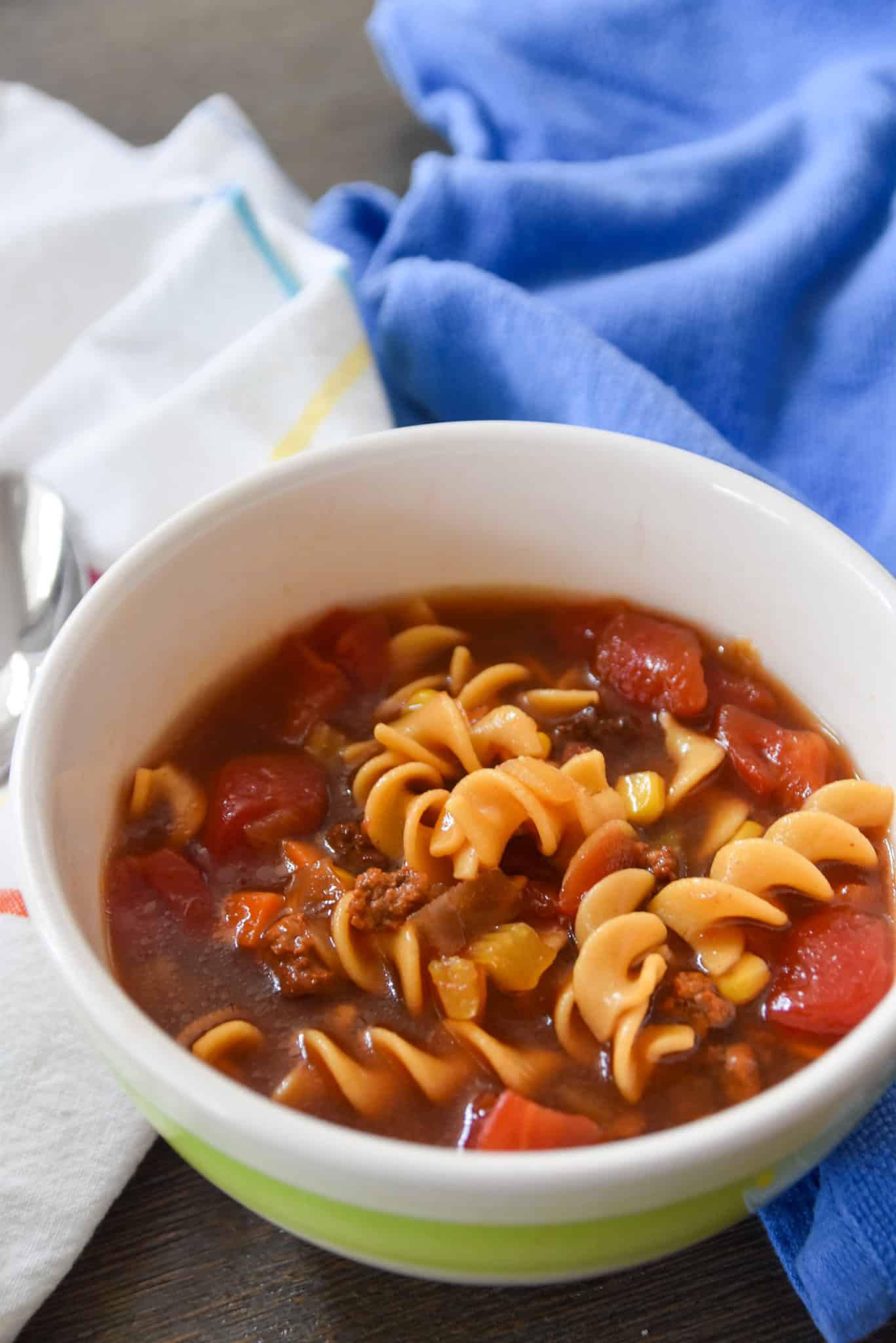 A bowl of tomato-based beef soup with rotini pasta and ground beef sits on a table near blue and white towels.