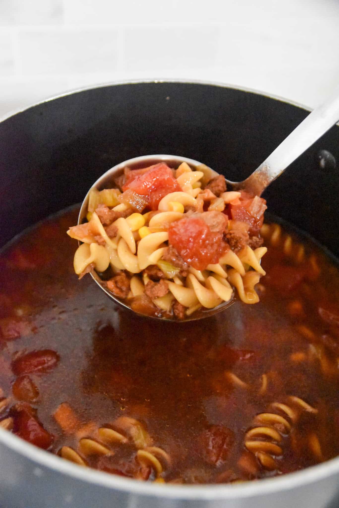 A ladle lifts rotini pasta, tomatoes, and tender beef from a pot of flavorful ground beef soup with red broth.