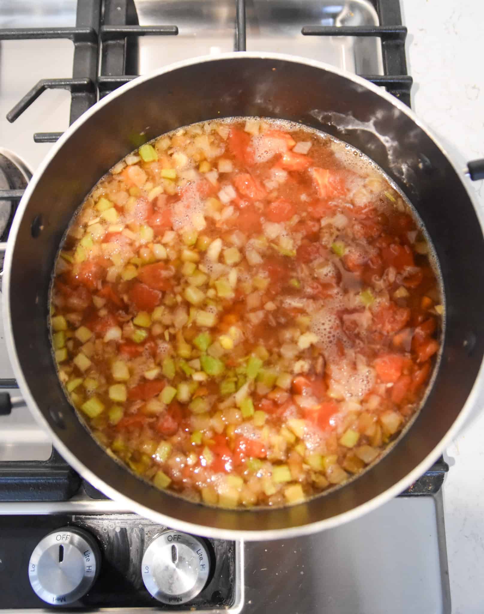 Diced vegetables and tomatoes simmering with ground beef in a pot on a gas stove for a hearty beef soup.