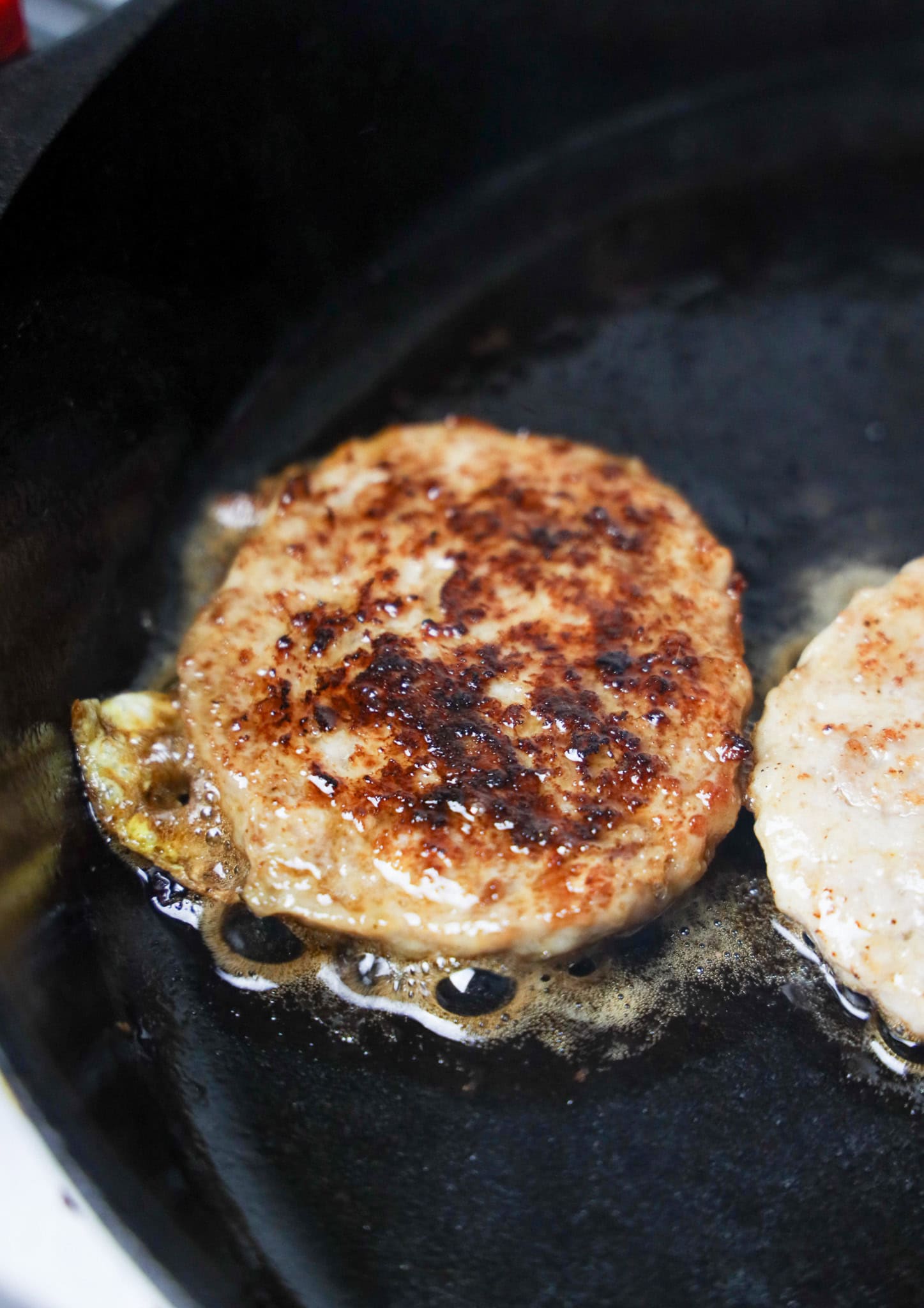 A browned burger patty cooking in a black skillet with sizzling juices around it, perfect for a homemade recipe inspired by your favorite Sausage Egg McMuffin.
